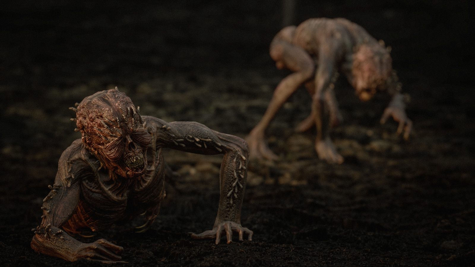 Two emaciated, skinless creatures on dark ground, one crawling towards the camera and one crouched behind it